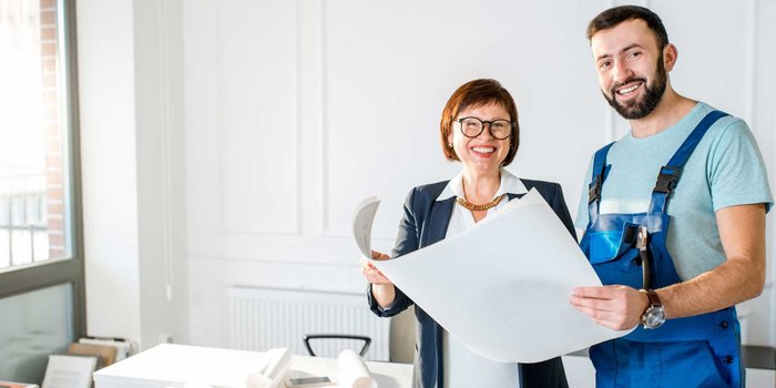 Ein Mann in Arbeitskleidung steht neben einer Frau, die eine Zeichnung in der Hand haelt