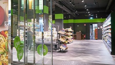Der Eingang eines modernen Supermarktes mit gläsernen Türen und bunten Regalen voller Lebensmittel ist sichtbar.